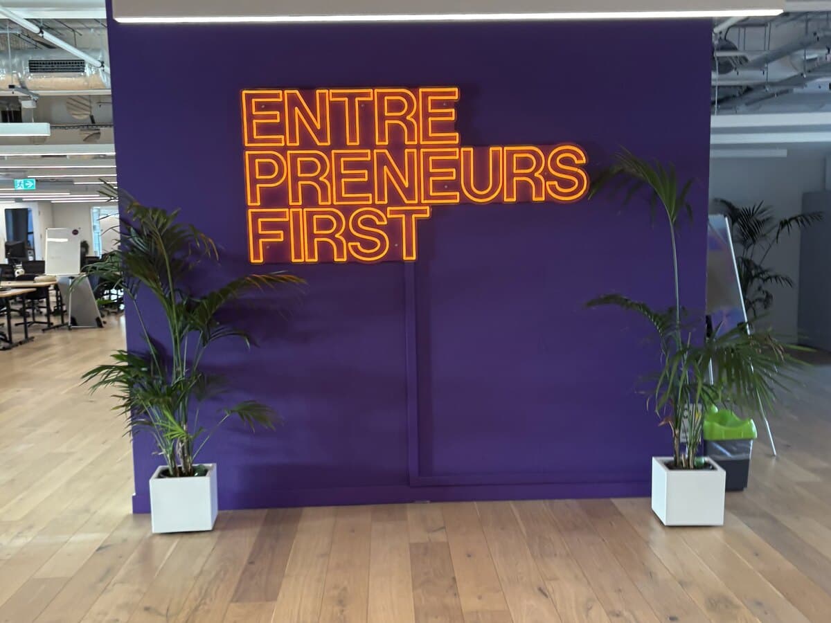 The Entrepreneurs First office in London: an orange neon sign reading ENTREPRENEURS FIRST mounted on a deep purple wall, with the open-plan floor visible on either side.