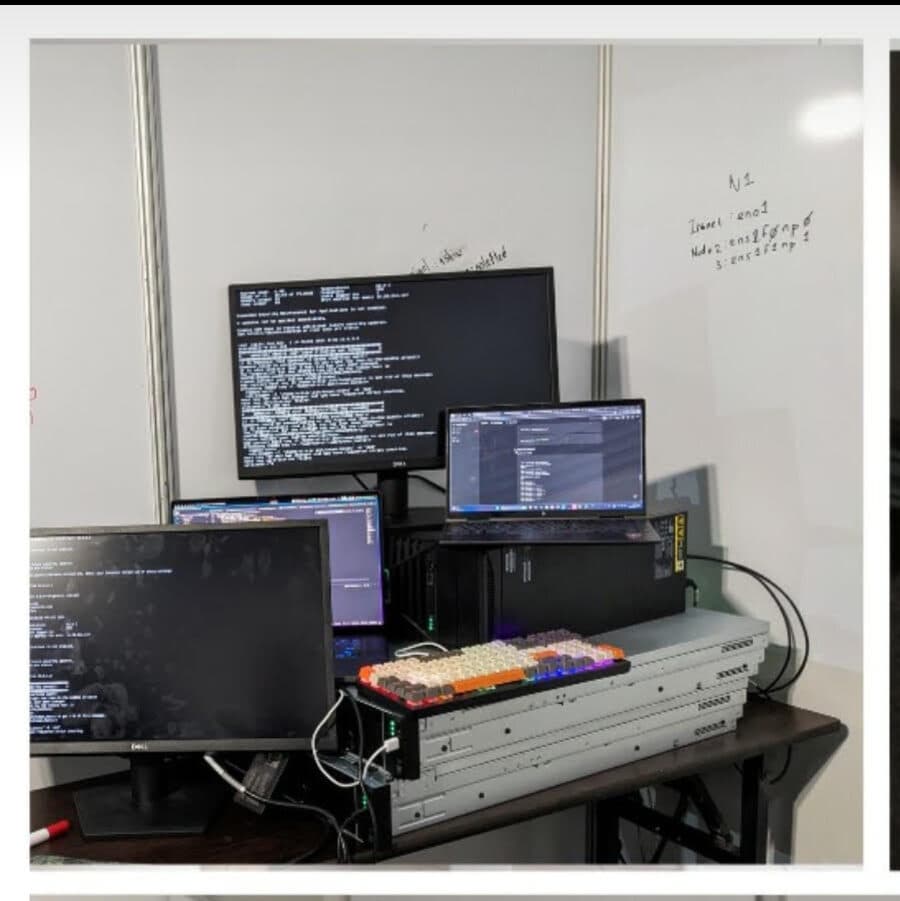The competition cluster: a rack server on a desk, three monitors of terminal output, and network interface names scrawled on the whiteboard behind it.